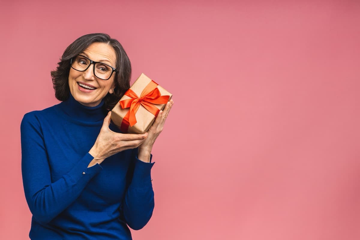 Happy woman holding wrapped birthday gift with red ribbon against pink background