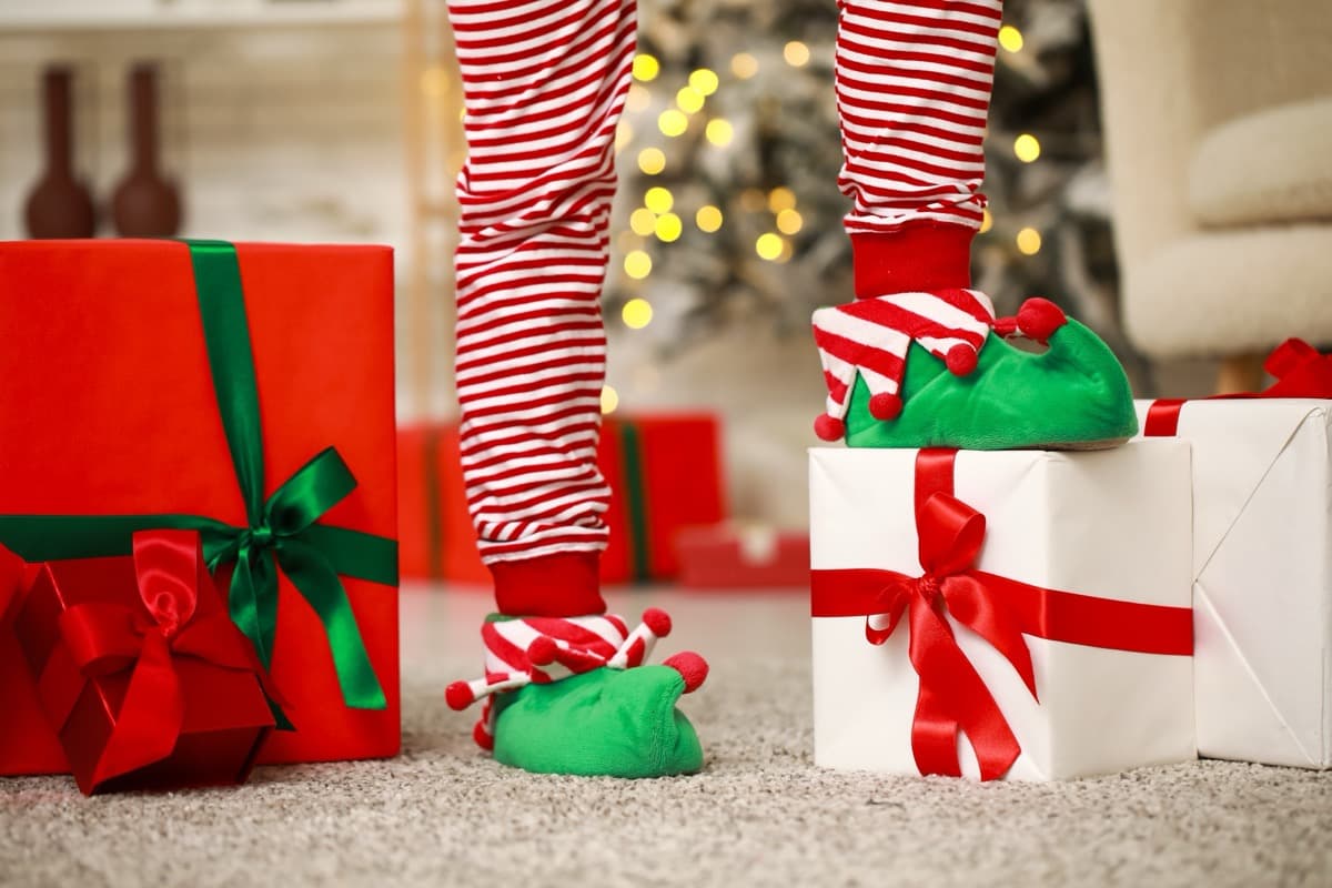 Child feet with elf slippers next to Christmas presents under tree